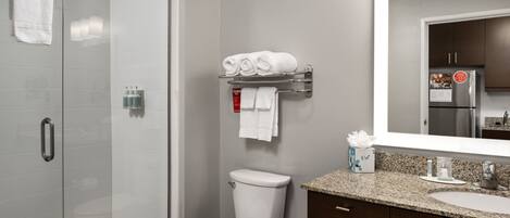 A modern bathroom with a glass shower enclosure, a toilet, a towel rack with neatly folded towels, a sink with a mirror, and a refrigerator in the background.