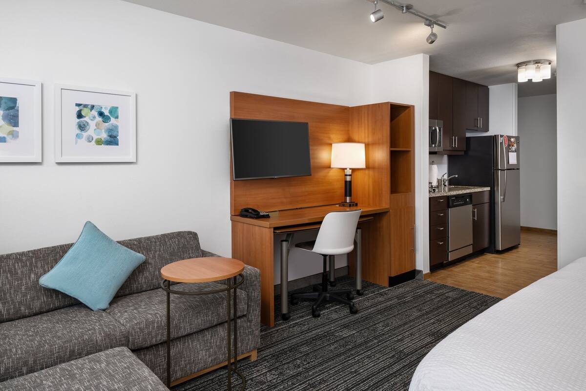 Une chambre d’hôtel moderne équipée d’un canapé, d’une petite table ronde, d’un bureau avec une chaise, d’une télévision et d’une kitchenette.