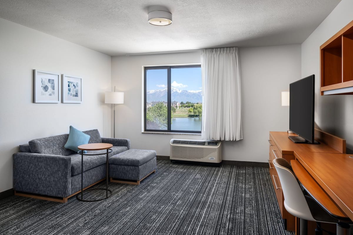 Une chambre d’hôtel moderne avec un canapé gris, une petite table ronde, un bureau avec une chaise, un téléviseur à écran plat, une grande fenêtre donnant sur les montagnes et deux tableaux encadrés accrochés au mur.