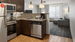 A modern kitchen with stainless steel appliances, a dishwasher, and granite countertops. There is a dining area with a gray sofa and a lamp. - Towneplace Suites Salt Lake City-West Valley (West Valley City)