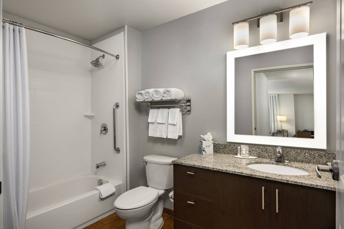 Une salle de bain moderne avec une baignoire, des toilettes et un lavabo. On y trouve un rideau de douche, un porte-serviettes avec des serviettes et un miroir avec des lumières au-dessus du lavabo.