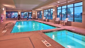 An indoor swimming pool with clear water, surrounded by lounge chairs and tables, large windows offering views of the outdoors, and a lifeguard chair. - Towneplace Suites Salt Lake City-West Valley (West Valley City)