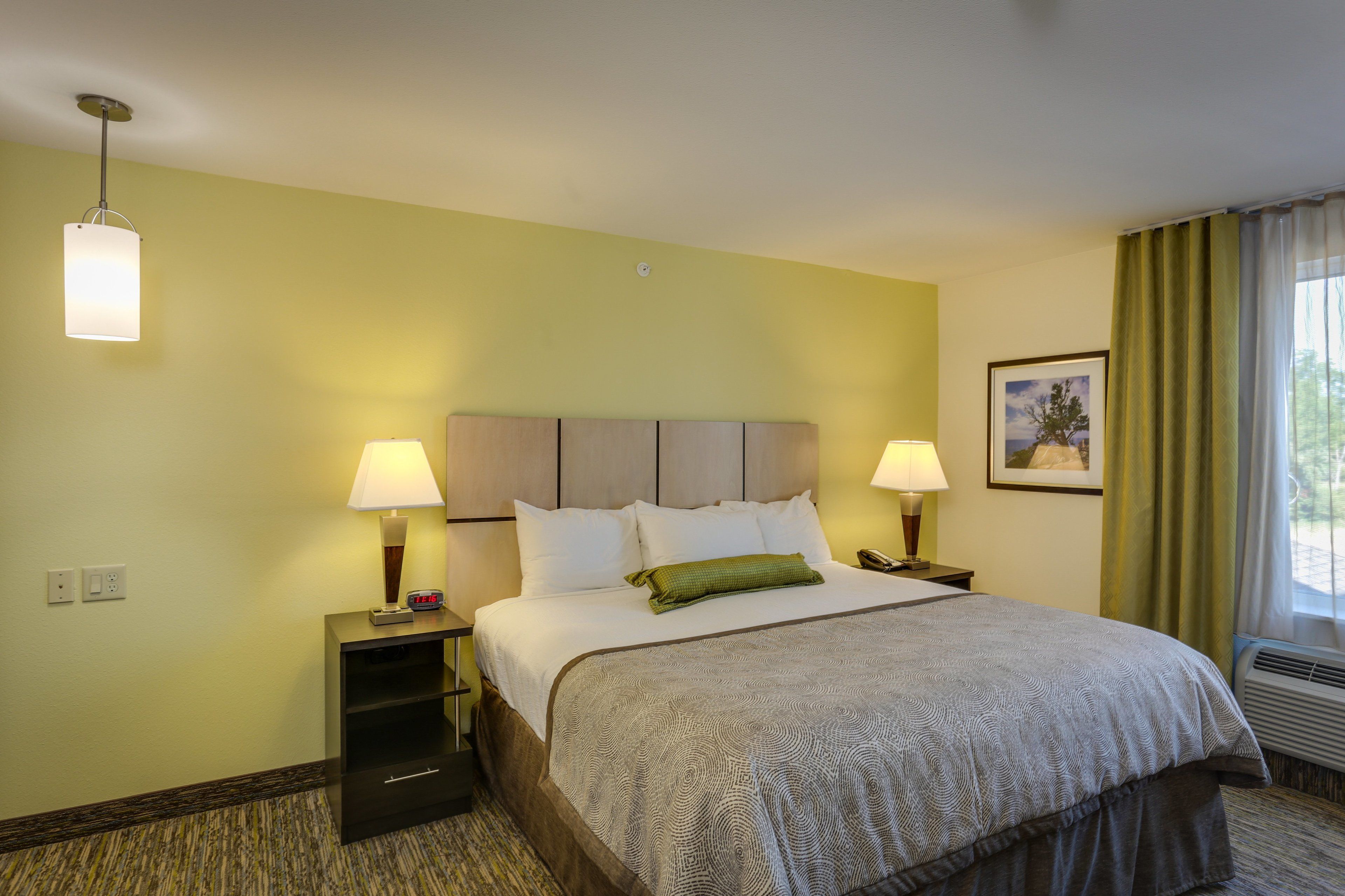 Quarto de hotel com cama, duas luminárias de cabeceira, mesa de cabeceira, quadro na parede e janela com cortinas.