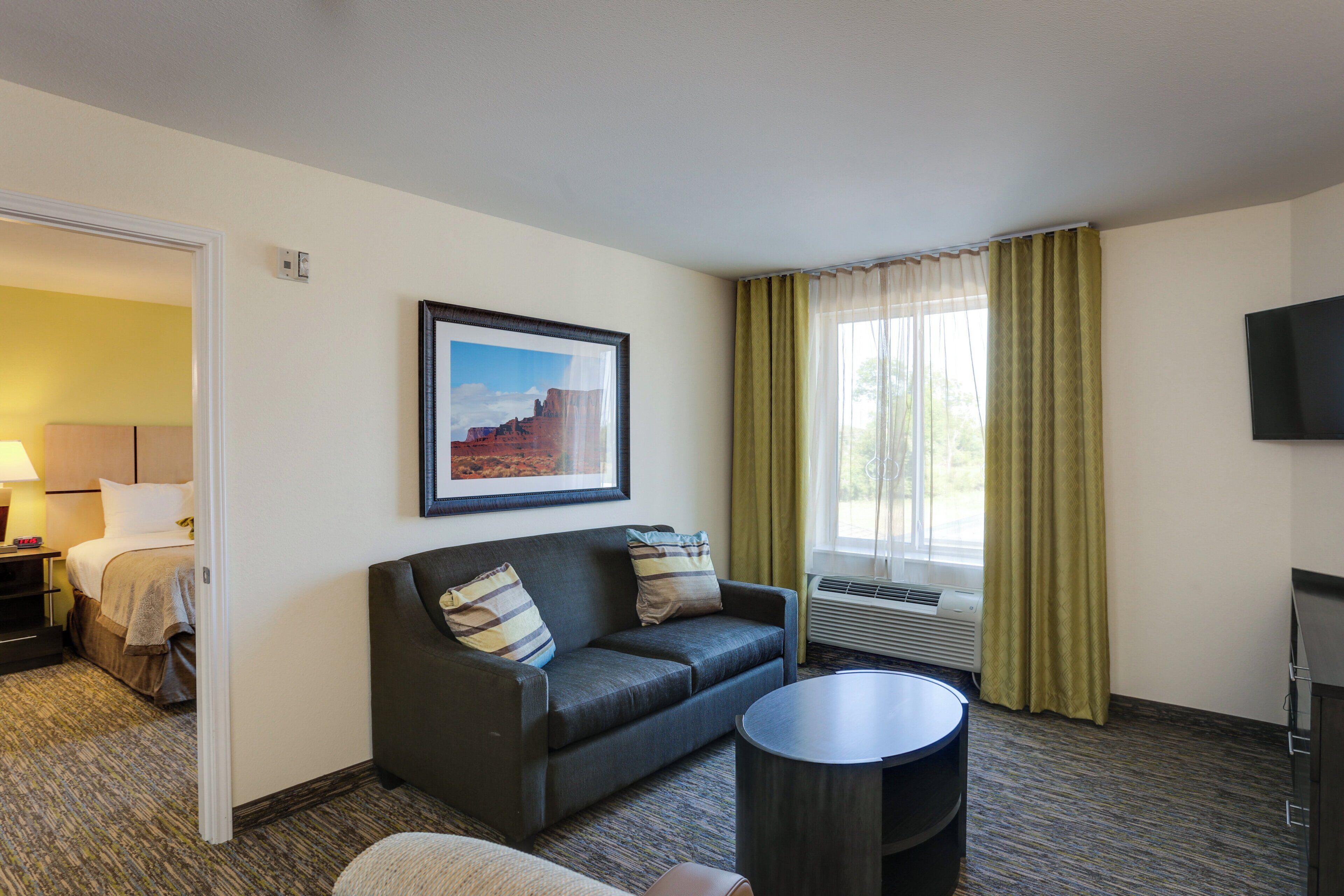 Habitación de hotel con cama, sofá, mesa de centro, televisor y ventana con cortinas.