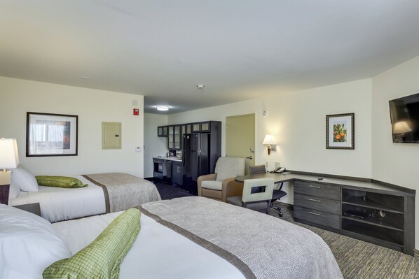 A hotel room with two beds, a desk, a chair, a TV, and a kitchenette.