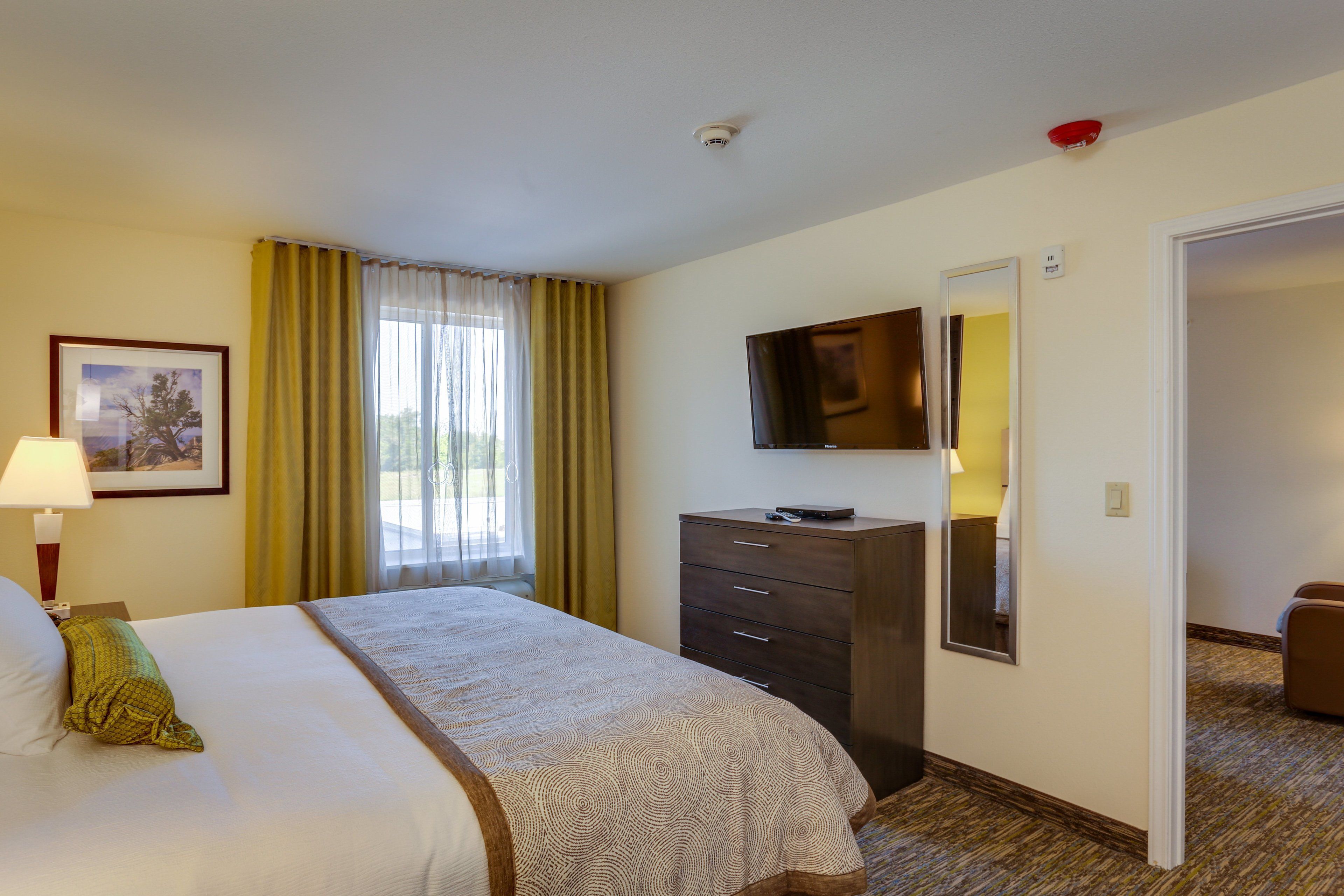 A hotel room with a bed, a dresser, a television mounted on the wall, and a door leading to another room.