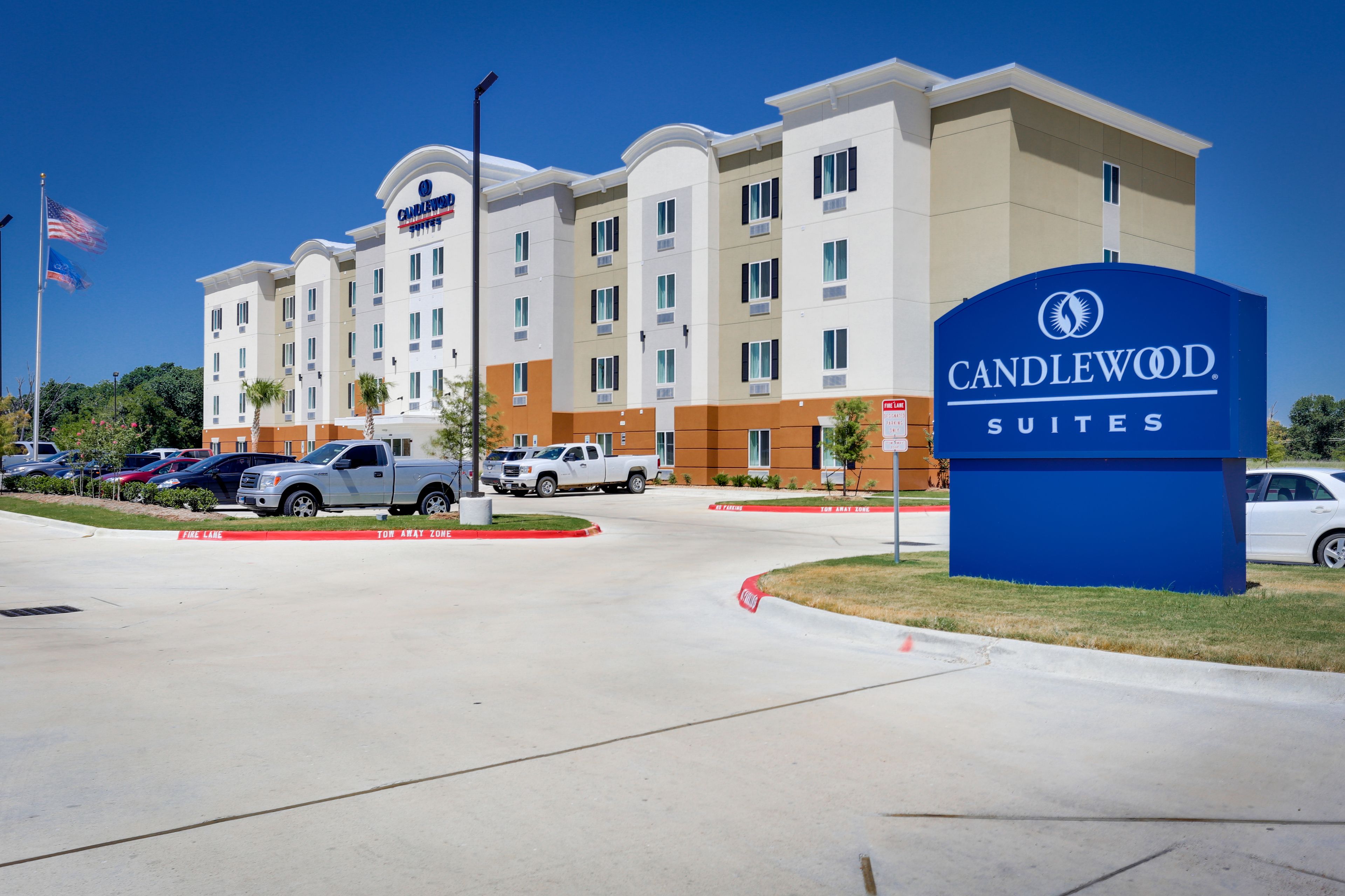 A hotel entrance with a sign for Candlewood Suites, a parking lot with several vehicles, and a clear blue sky.