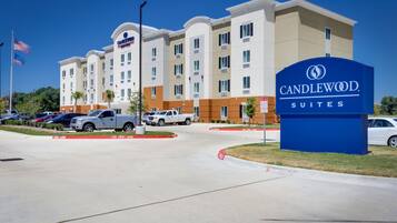 A hotel entrance with a sign for Candlewood Suites, a parking lot with several vehicles, and a clear blue sky.