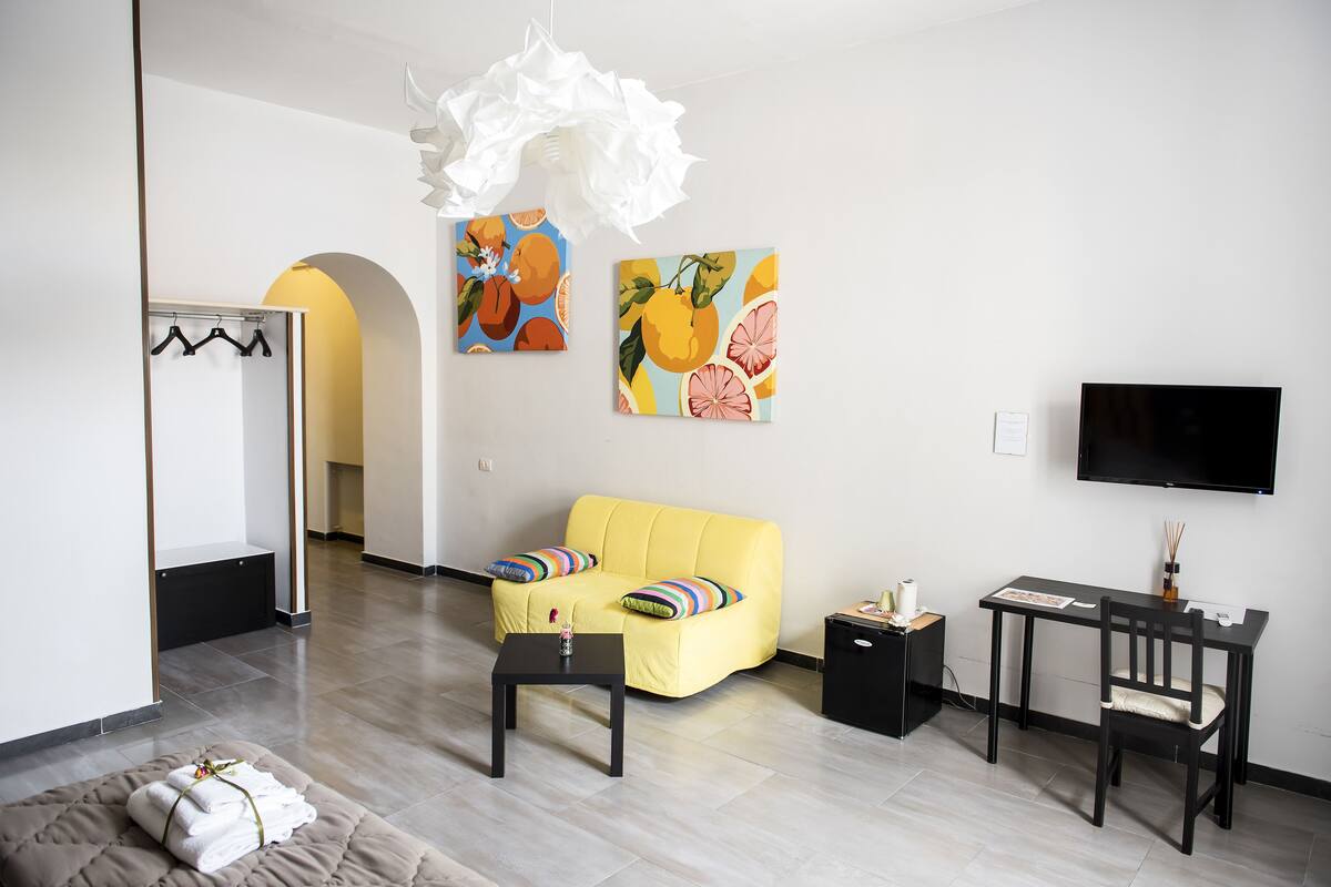 Un salon moderne avec un canapé jaune, une table noire et un téléviseur. Deux tableaux ornent le mur, ainsi qu’un meuble noir et une porte blanche avec un porte-manteau.