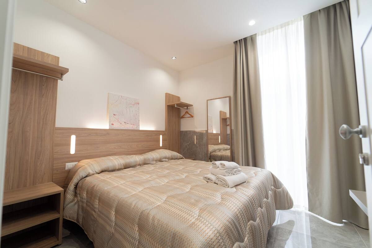 Une chambre d’hôtel avec un grand lit, une tête de lit en bois, une table de chevet, un miroir et une fenêtre avec des rideaux.