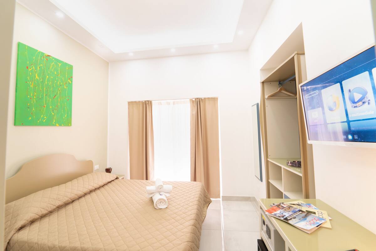 Une chambre d’hôtel avec un lit, une télévision, un bureau avec un magazine, et un tableau abstrait vert accroché au mur.
