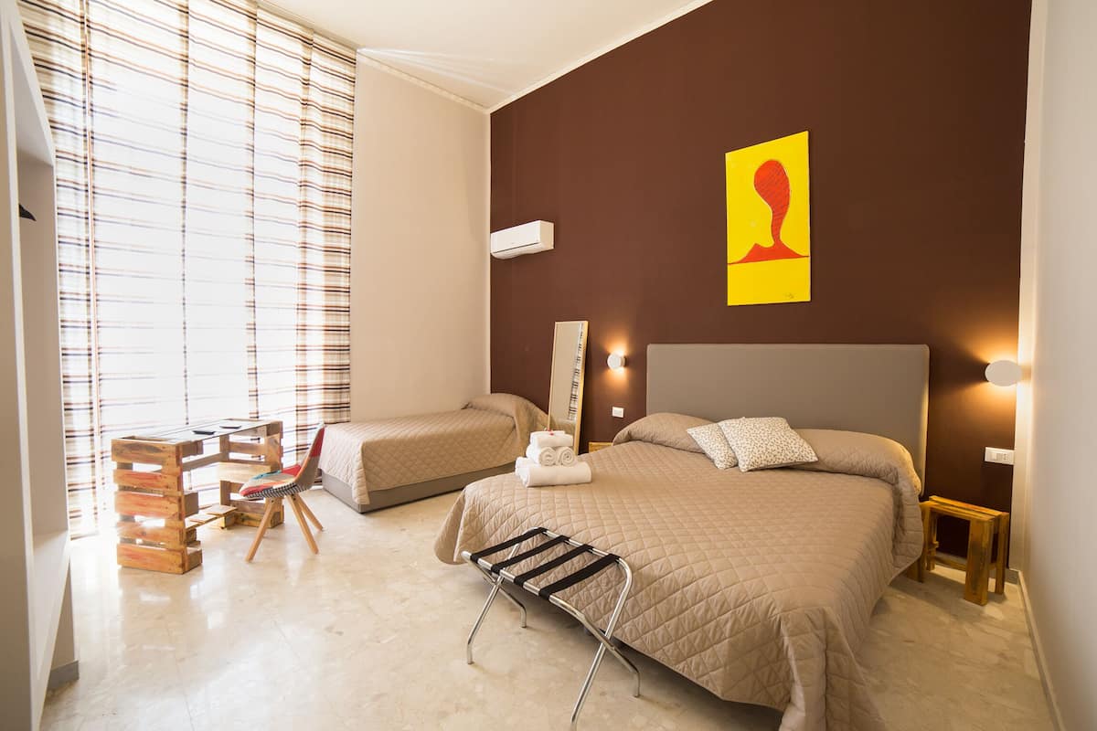 Une chambre d’hôtel avec deux lits, un bureau en bois, une chaise et une fenêtre avec des stores.