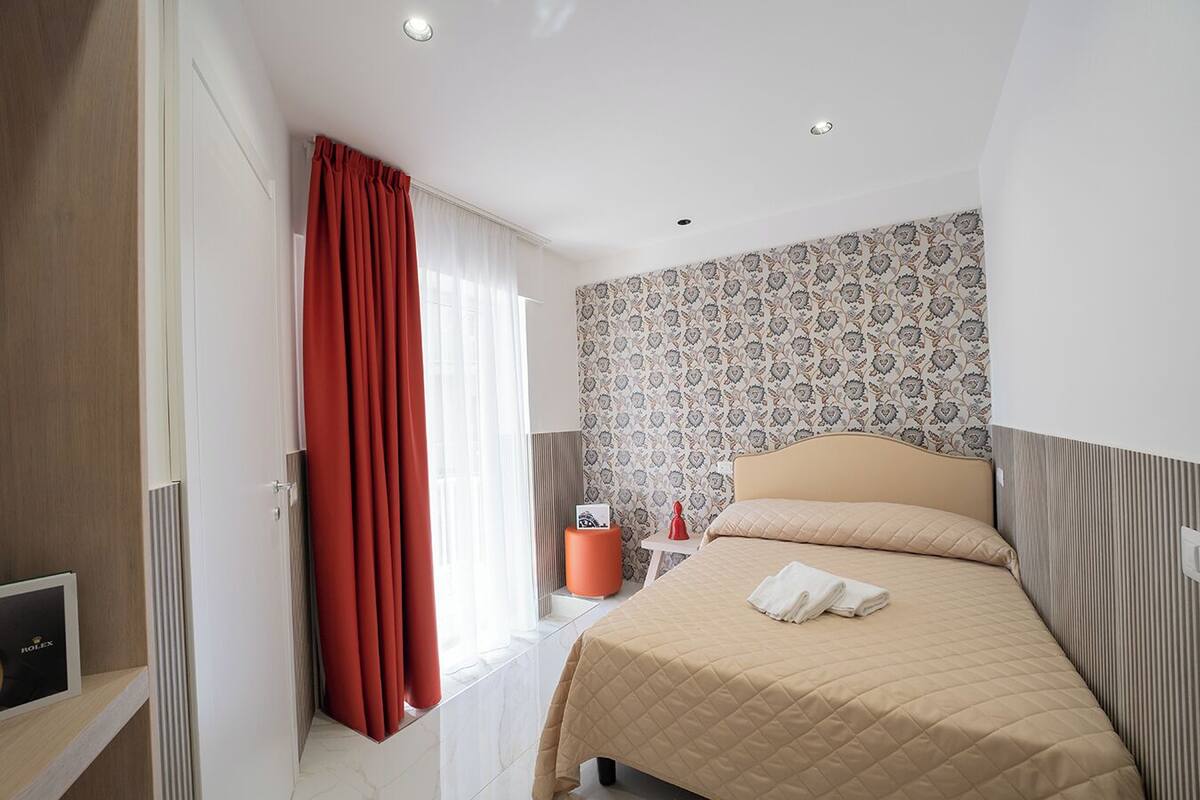 Une chambre d’hôtel avec un lit, une table de chevet, un rideau rouge, un papier peint à motifs et une fenêtre avec des rideaux blancs.