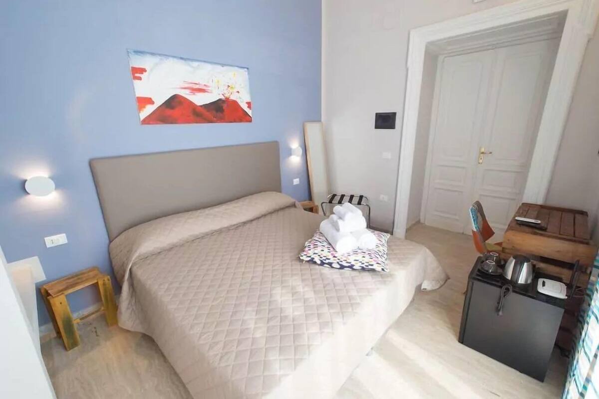 Une chambre à coucher avec un lit, un petit tabouret en bois, une table de chevet avec une bouilloire et une porte.