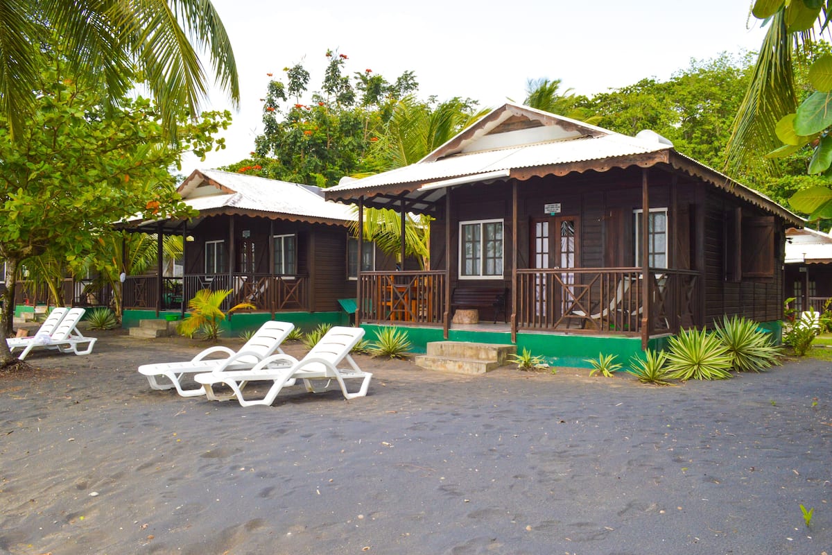 A wooden bungalow with a porch, surrounded by palm trees and a sandy area with white lounge chairs.