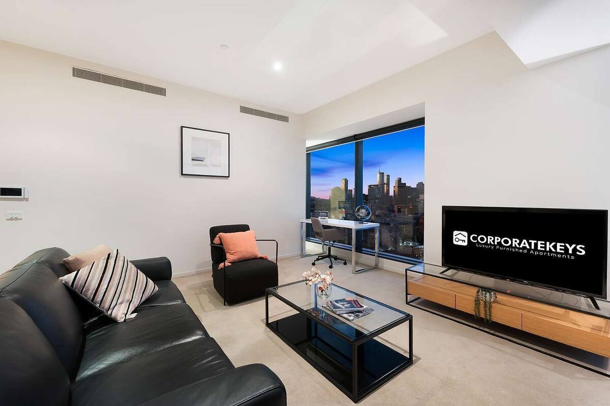 A modern living room with a black leather sofa, a glass coffee table, a black office chair, a large TV, and a city view through large windows.