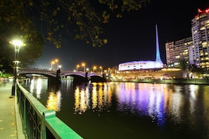 A nighttime view of a riverside area with a bridge, a lit-up building with a spire, and a restaurant with outdoor seating.