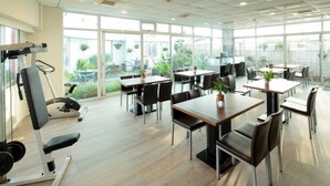 Un vaste espace intérieur doté de nombreuses tables et chaises, une salle de sport équipée de matériel d’exercice et de grandes fenêtres offrant une vue sur l’extérieur.