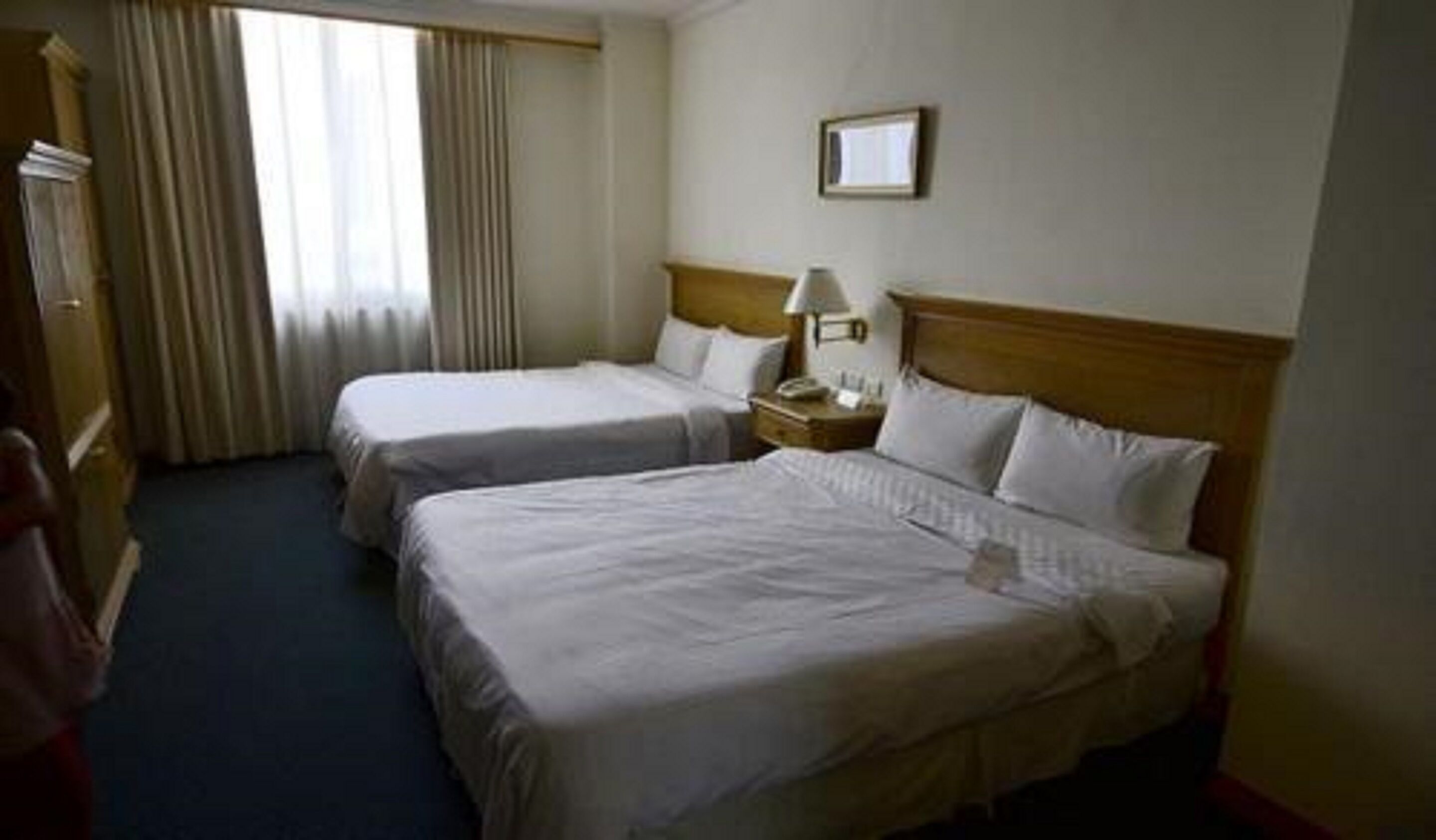 Habitación de hotel con dos camas, cabecero de madera, mesita de noche con lámpara y ventana con cortinas.