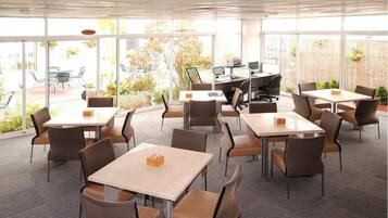 A modern indoor dining area with multiple tables and chairs, large windows, and a view of the outside garden.