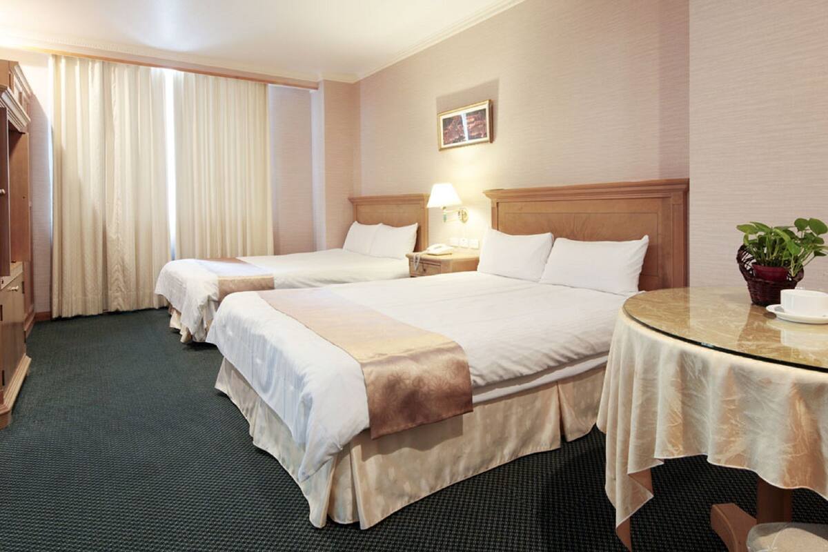 A hotel room with two beds, a round table with a teapot and cup, a nightstand with a lamp, and a potted plant.