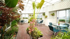 Un patio abrité, doté de tables et de chaises, entouré de différentes plantes et surmonté d’un toit en verre.