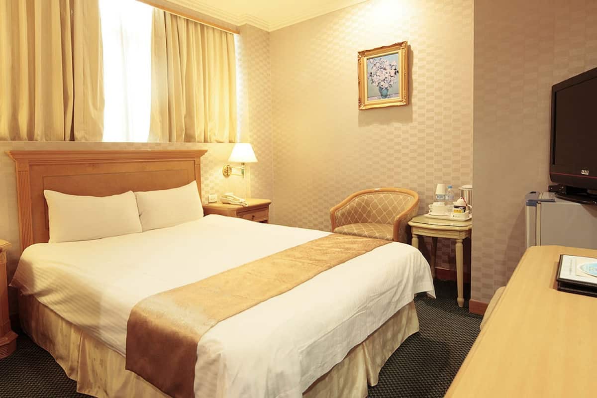 A hotel room with a large bed, two bedside tables with lamps, a chair, a desk with a television, and a framed picture on the wall.