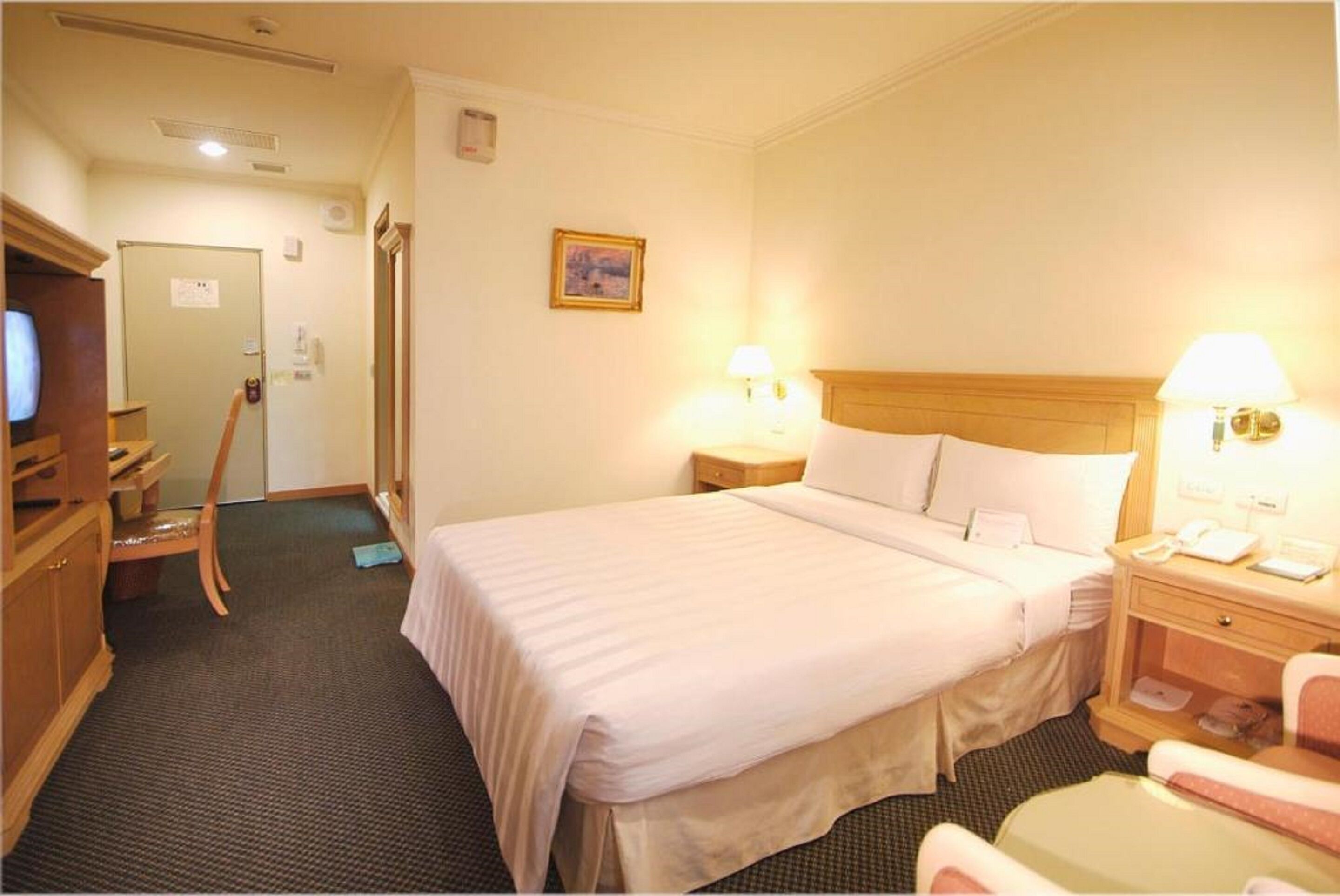 Habitació d'hotel amb un llit de matrimoni, taules de nit amb làmpades, un armari de fusta, un escriptori amb una cadira i una televisió.