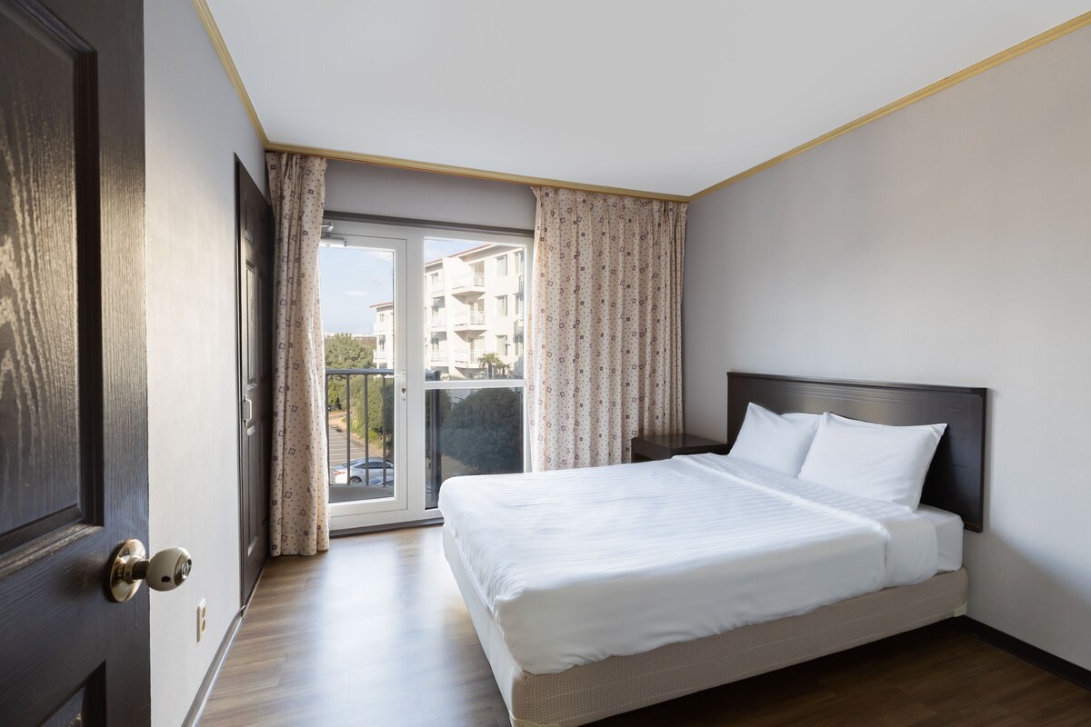 A hotel room with a bed, a door, and a view through a glass door to the outside.