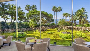 A modern interior with large windows, a seating area with chairs and a table, and a view of a landscaped garden with palm trees and a stone wall. - Kensington Resort Seogwipo (Seogwipo)