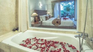 A hotel room with a large bathtub filled with rose petals, a bed with a patterned blanket, and a view of a balcony with trees. - Gita Maha Hotel (Ubud)