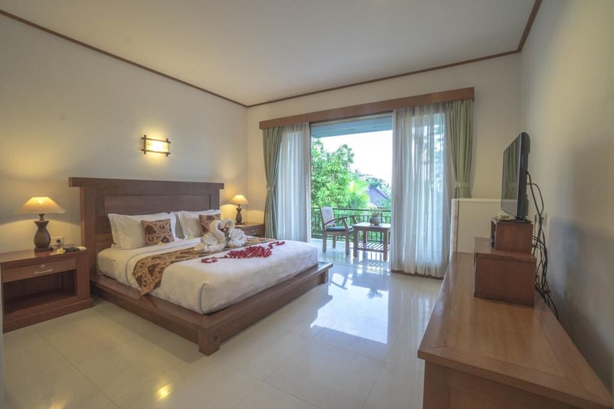 A hotel room with a large bed, two bedside tables with lamps, a wooden desk, a television, and a balcony with a table and chairs.