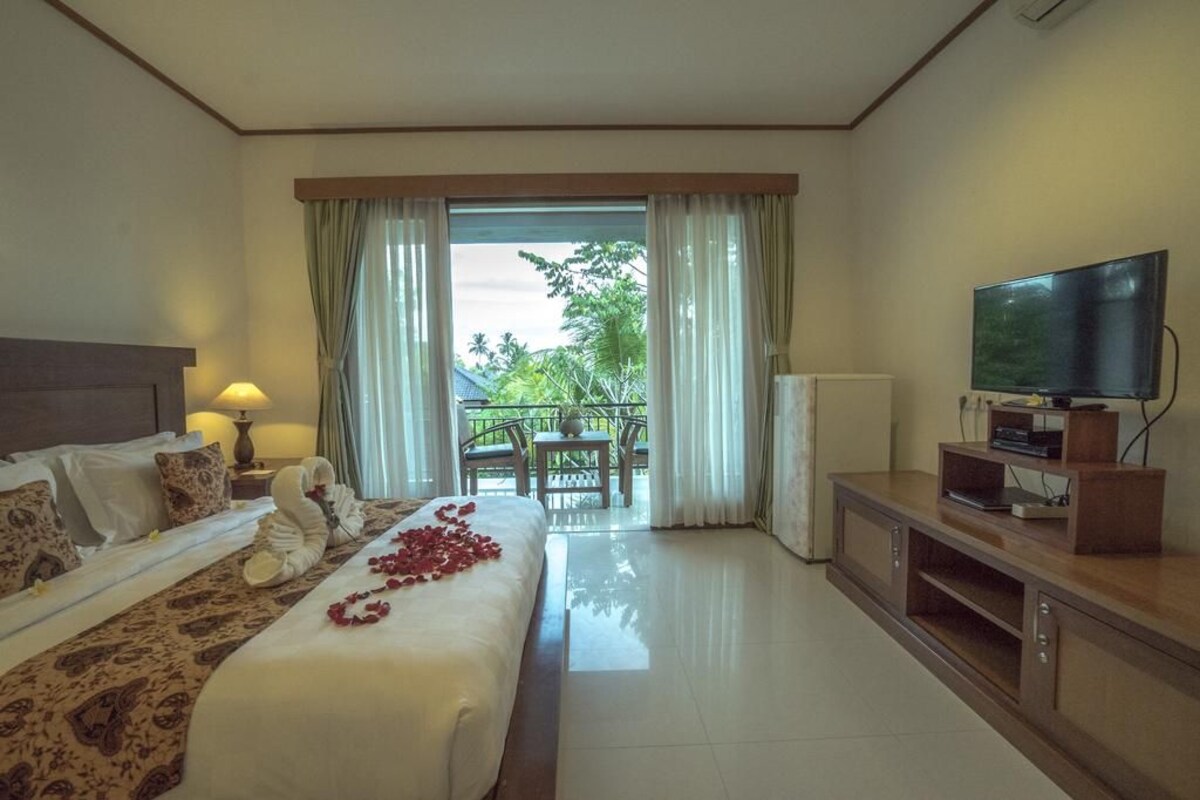 A hotel room with a bed, a television, a balcony with a view of greenery, and a table with chairs.