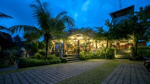 A well-lit hotel entrance with a paved pathway leading to the main building, surrounded by lush greenery and palm trees. - Gita Maha Hotel (Ubud)