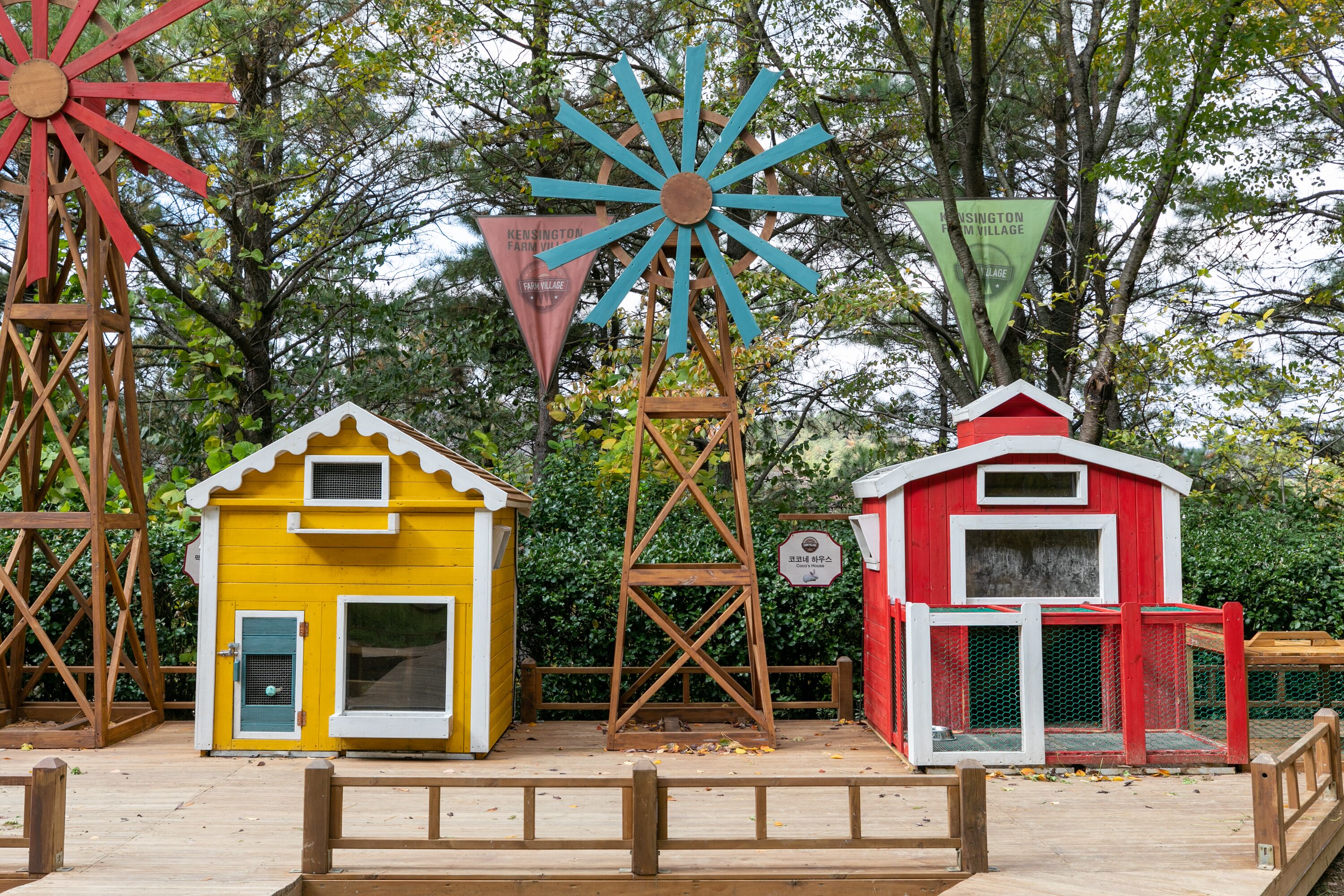 A wooden playhouse with a yellow exterior and a red roof, a blue windmill, and a red barn-like structure.