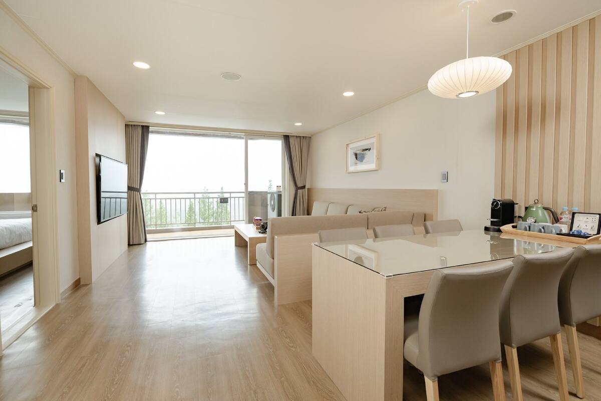 A modern hotel room with a glass dining table, a sofa, a bed, and a balcony with curtains.