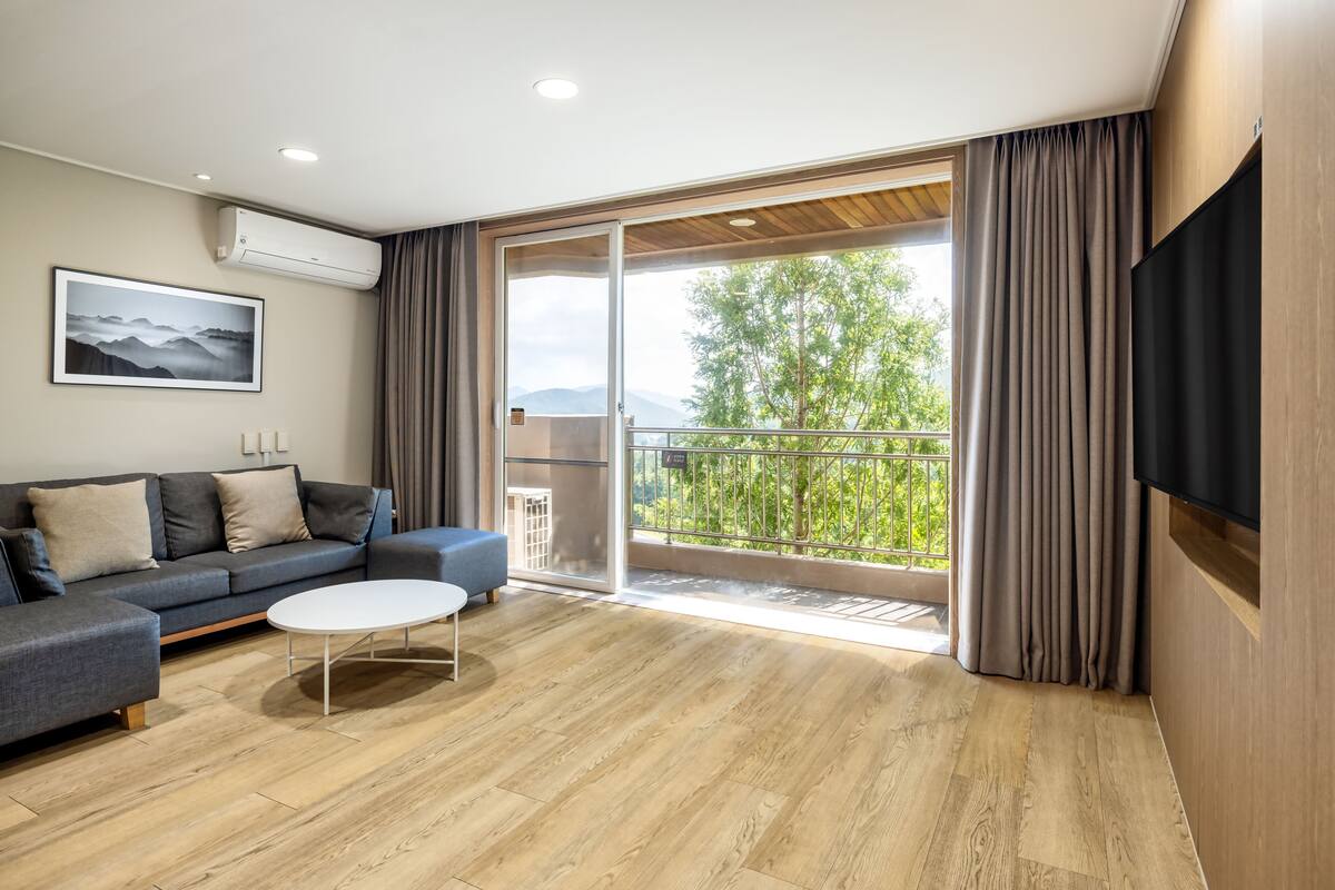 Ein modernes Wohnzimmer mit grauem Sofa, einem weißen Couchtisch und einer großen Schiebeglasdoor zum Balkon mit Blick auf Bäume und Berge.