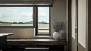 Ein Zimmer mit einem großen Fenster, einer weißen Vase mit Zweigen und einer hölzernen Bank.