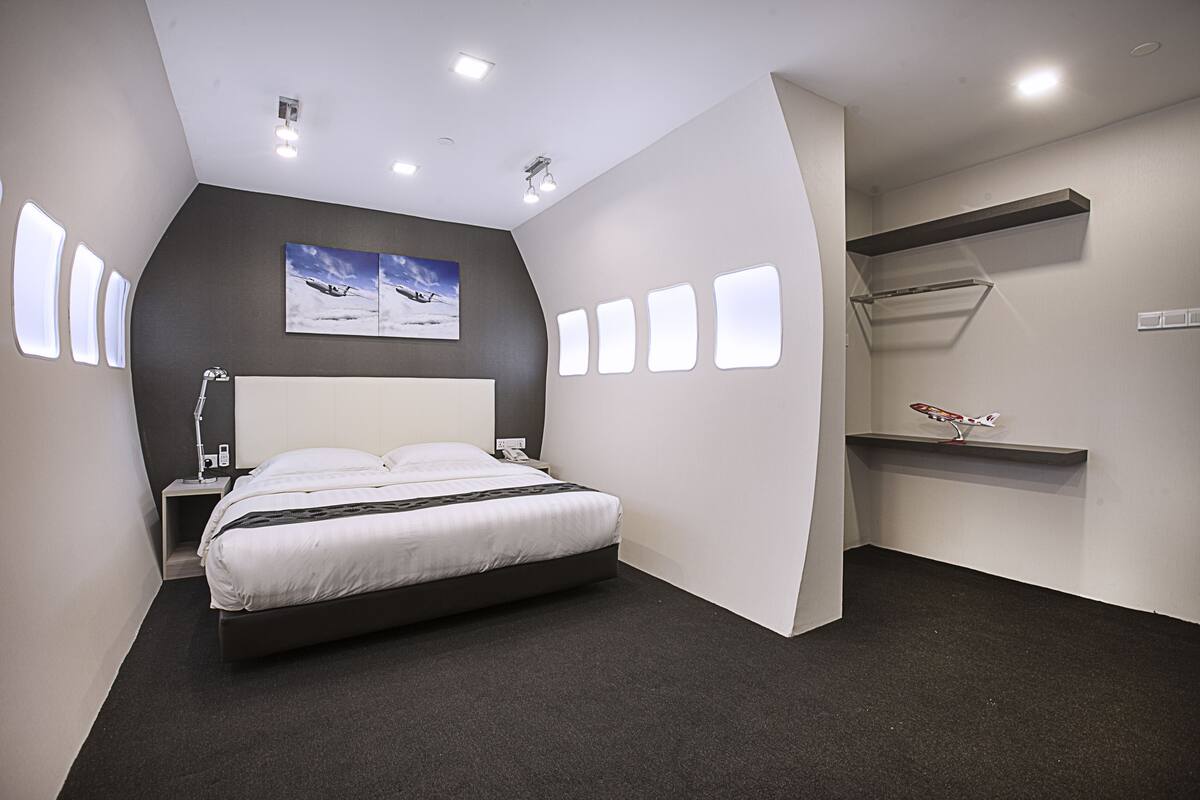 A modern hotel room with a large bed, a desk, and a chair. There are multiple windows and a ceiling light fixture.