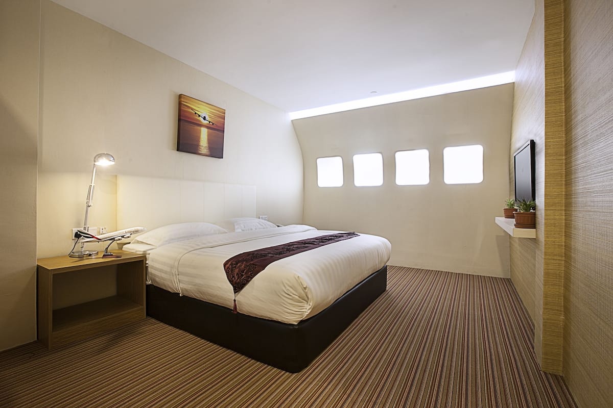 A hotel room with a bed, bedside table, lamp, artwork, windows, and a TV on a shelf.