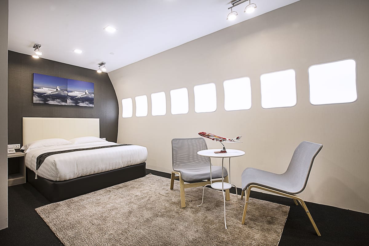 A modern hotel room with a bed, two chairs, a small table, and a wall with illuminated windows.