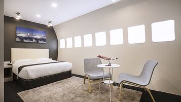 Quarto de hotel moderno com uma cama, duas cadeiras, uma mesa pequena e uma parede com janelas iluminadas.