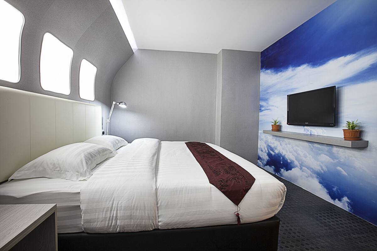 A modern hotel room with a large bed, a mounted TV, and a wall mural of clouds.