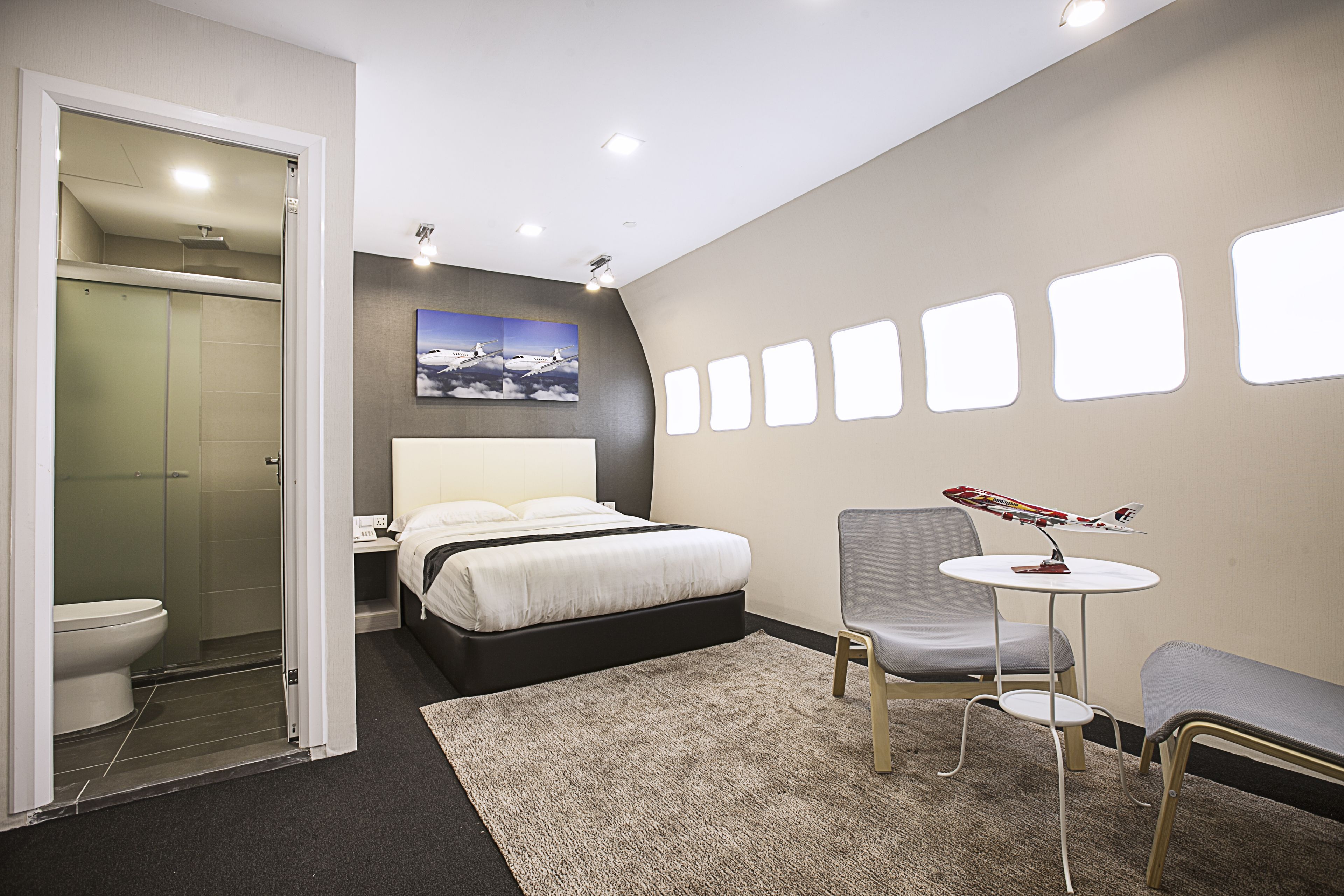 A modern hotel room with a bed, a small table with a model airplane, a chair, a shower, and a toilet.