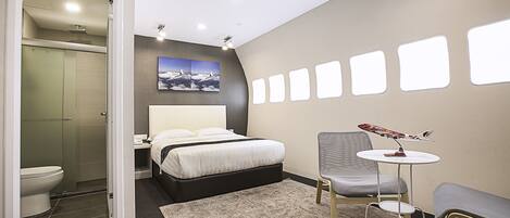 A modern hotel room with a bed, a small table with a model airplane, a chair, a shower, and a toilet.
