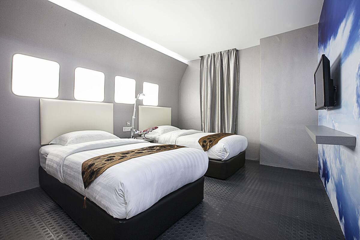 A hotel room with two beds, a bedside table, a lamp, a TV, and a wall-mounted shelf.