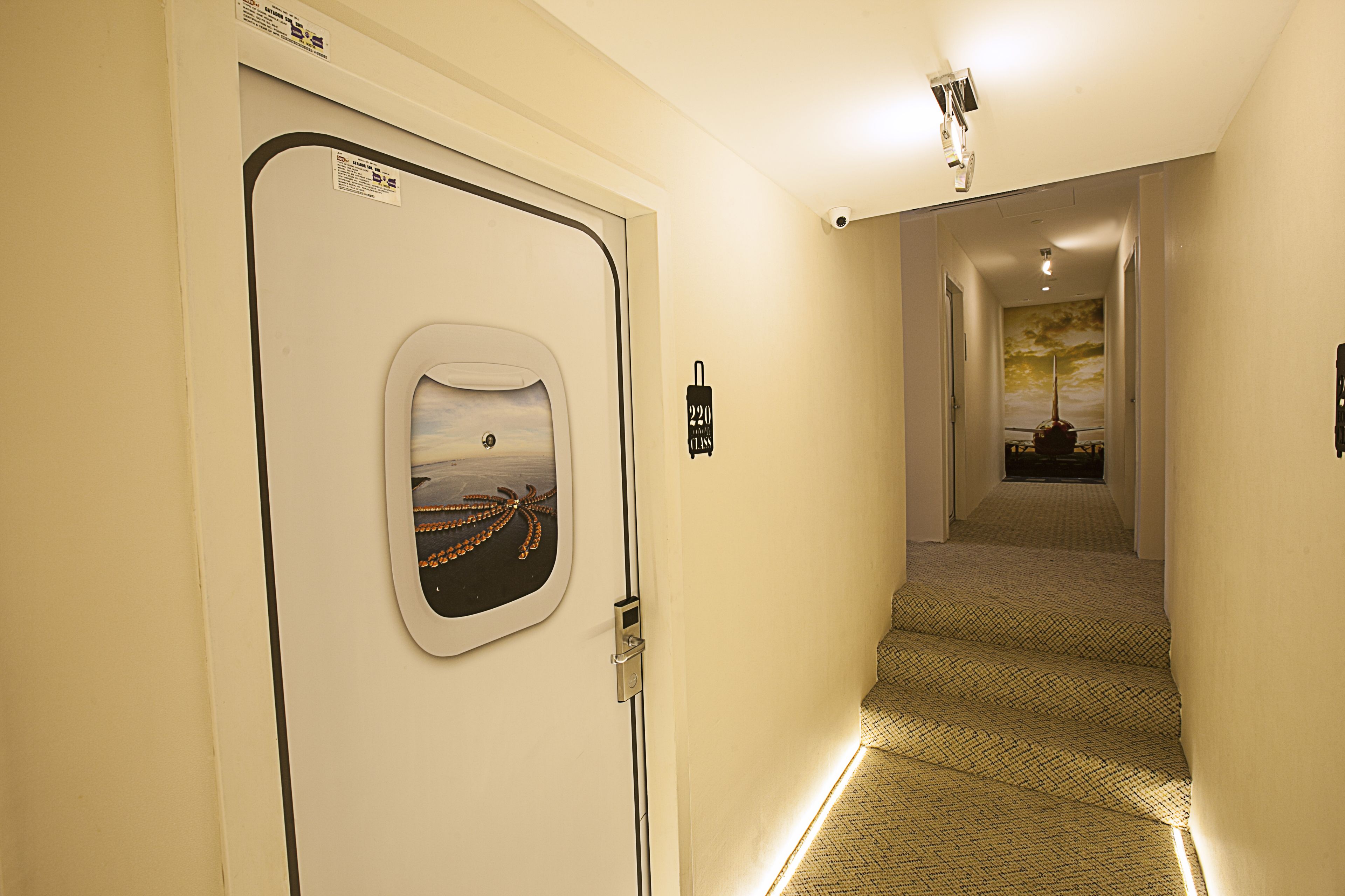 Um corredor de hotel com escadas revestidas de carpete, uma porta com janela e uma câmera de segurança.