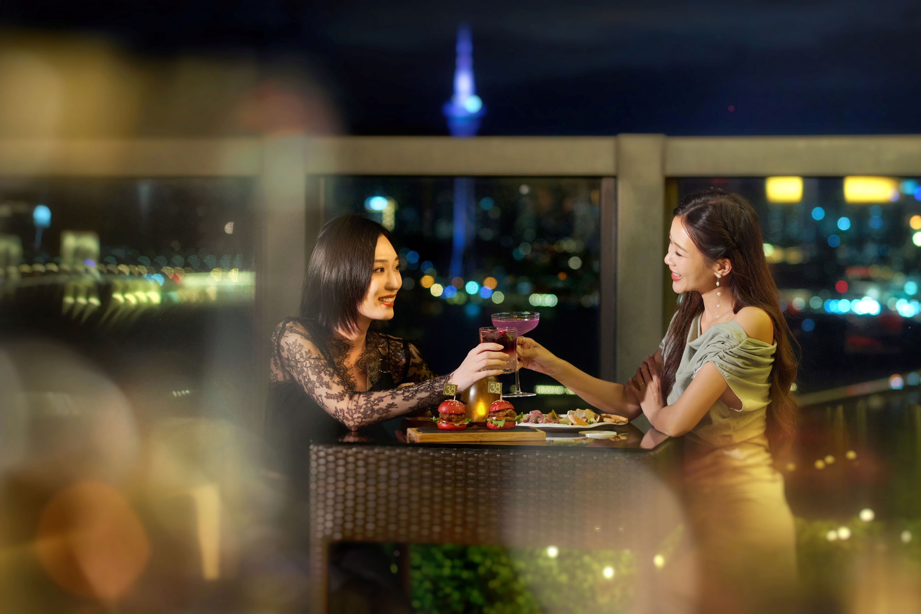 Two people sitting at a table with drinks and burgers, overlooking a cityscape with a tower in the background.