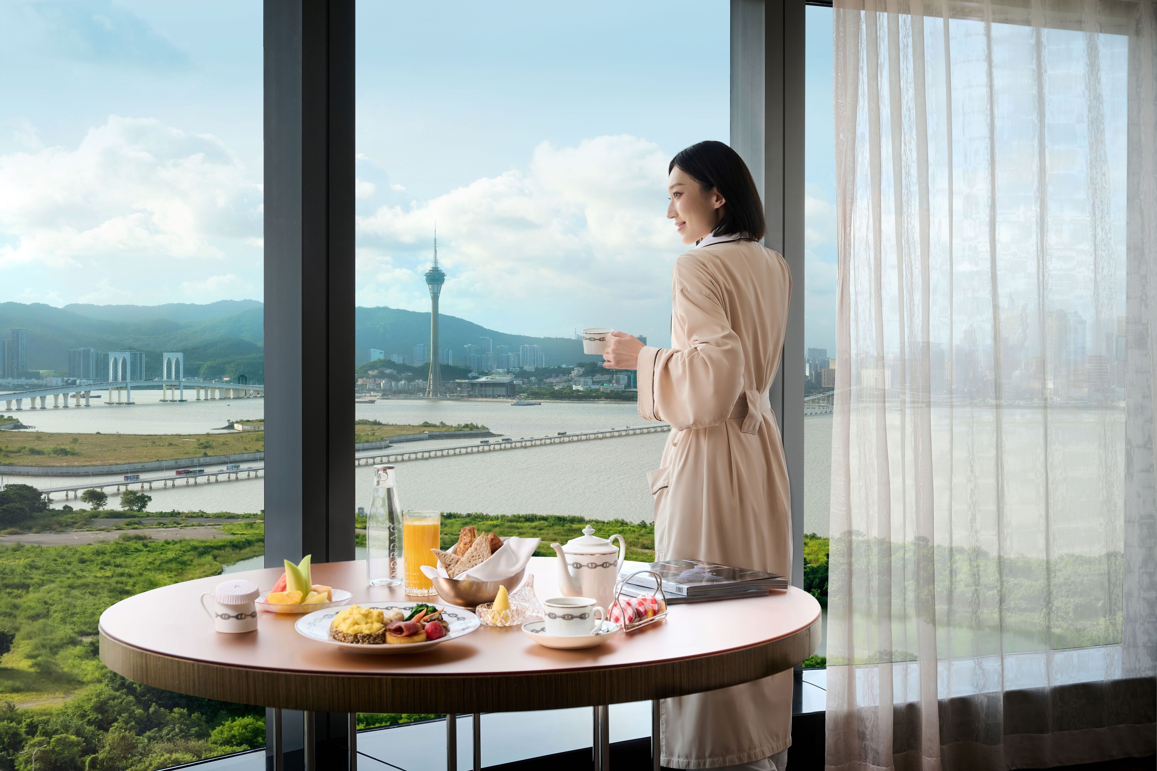 Una habitación con un amplio ventanal que da a un paisaje urbano, una mesa con alimentos para el desayuno y una persona de pie junto a la ventana.