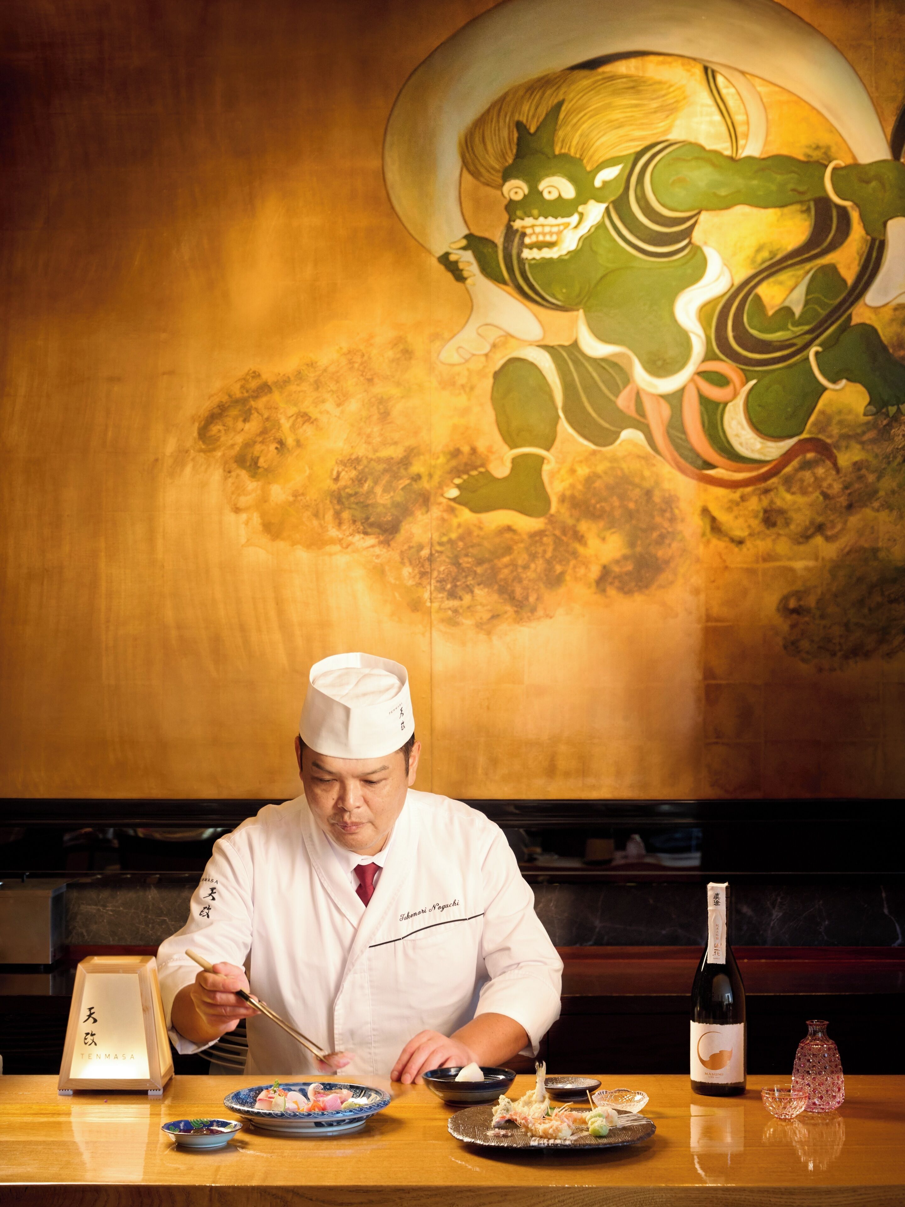 A chef in a white uniform preparing food at a wooden table with a traditional Japanese backdrop.